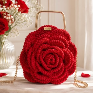 Può includere: Una borsa all'uncinetto rosso vivace a forma di rosa in fiore, dotata di un manico superiore in metallo dorato e di una tracolla a catena dorata abbinata. Una piccola etichetta rettangolare dorata sul davanti riporta il testo Halmi Mariana Art.Cros.