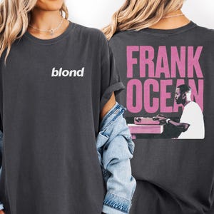 a woman wearing a black t-shirt with the text "blond" and a graphic of a man sitting at a desk. She is also wearing a denim jacket and has her arms crossed.