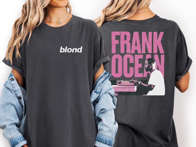 a woman wearing a black t-shirt with the text "blond" and a graphic of a man sitting at a desk. She is also wearing a denim jacket and has her arms crossed.