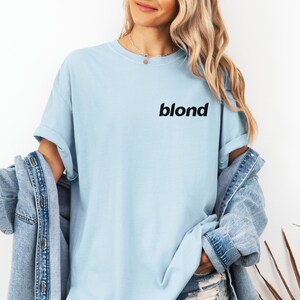A woman wearing a light blue t-shirt with the word "blond" printed on it, standing against a white background. She is also wearing a denim jacket and has her hands in her pockets.