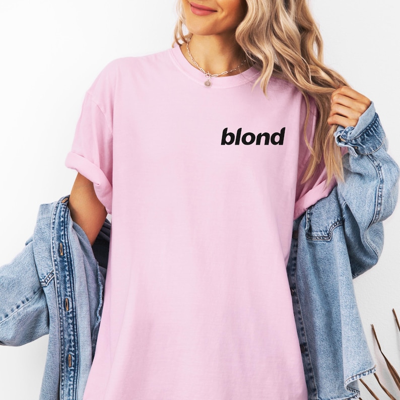 A woman wearing a pink t-shirt with the word "blond" printed on it, standing against a white background. She is also wearing a denim jacket and has her hair styled in loose waves.