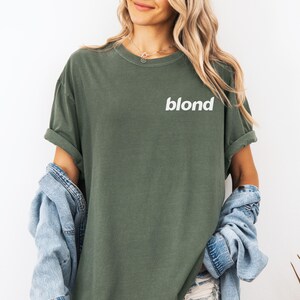 A woman wearing a green t-shirt with the word "blond" printed on it, standing against a white background.