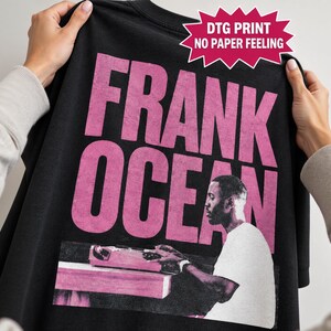 May include: A black t-shirt featuring large, distressed pink text that reads FRANK OCEAN. Below the text is a monochromatic graphic of a person sitting at a desk with a typewriter. A pink starburst graphic includes the text DTG PRINT NO PAPER FEELING.