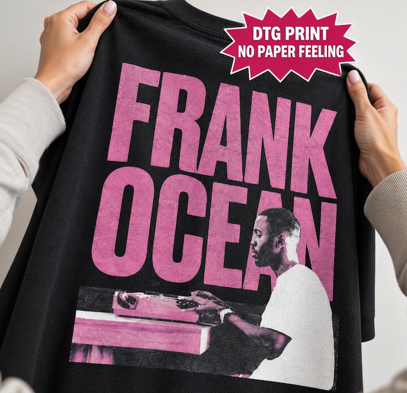 May include: A black t-shirt featuring large, distressed pink text that reads FRANK OCEAN. Below the text is a monochromatic graphic of a person sitting at a desk with a typewriter. A pink starburst graphic includes the text DTG PRINT NO PAPER FEELING.