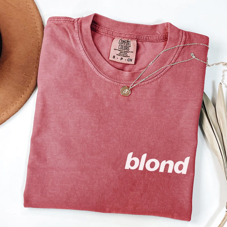 A red t-shirt with the word "blond" printed on it, along with a necklace and a brown hat, are displayed on a white surface.