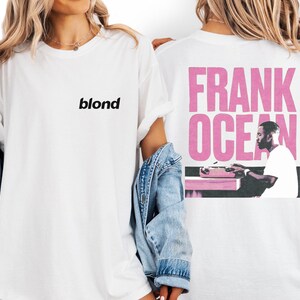 A woman wearing a white t-shirt with the word "blond" printed on it, standing next to a denim jacket.