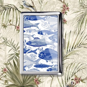 Puede incluir: Un estuche de cigarrillos rectangular, con marco plateado, que presenta un patrón de peces azul y blanco. El diseño incluye varias ilustraciones de peces sobre un fondo blanco. El estuche se encuentra sobre un fondo de follaje tropical.