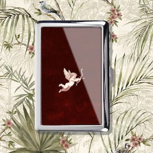 May include: A silver metal cigarette case with a burgundy interior featuring a cherub with a bow and arrow. The case has a rectangular shape and a shiny finish. The background is a floral pattern with birds and leaves.