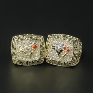 Può includere: Due anelli da campionato dorati con la scritta "WORLD CHAMPIONS". Ogni anello presenta una grande gemma trasparente circondata da pietre più piccole e un logo della squadra con dettagli rossi e blu.