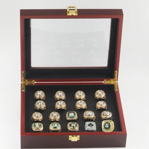May include: A wooden display case with a glass top, open to reveal multiple gold-toned rings with various designs and gemstones. The case has a dark red finish and a black interior, showcasing the rings.