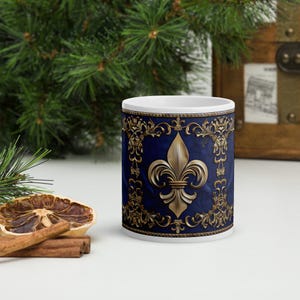 Może przedstawiać: Biały ceramiczny kubek z granatowym tłem i złotym wzorem fleur-de-lis. Kubek ma bogate złote zawijasy i złotą fleur-de-lis pośrodku. Kubek jest przedmiotem dekoracyjnym.