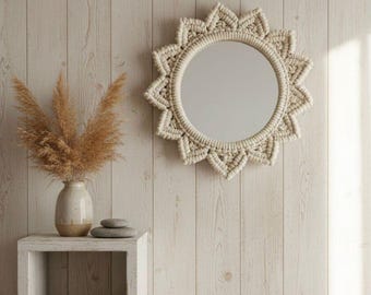 May include: A round mirror with a cream-colored macrame sunburst frame hangs on a light wood-paneled wall. A small white shelf holds a vase with dried plants, two gray stones, and a folded cream-colored blanket. The overall aesthetic is bohemian and natural.