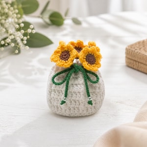 Peut inclure: Un petit vase crocheté gris clair, orné de trois tournesols jaunes et bruns. Le vase est noué avec une ficelle verte et comporte de petites perles argentées. L'arrangement est sur une surface blanche.