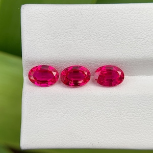 Könnte beinhalten: Drei ovale, leuchtend rote Edelsteine ​​werden auf einer weißen Oberfläche präsentiert. Die Edelsteine ​​sind gleichmäßig verteilt und scheinen poliert zu sein und Licht zu reflektieren. Der Hintergrund ist verschwommen, mit grünem Laub sichtbar.