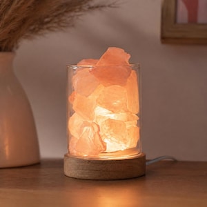 May include: A Himalayan salt lamp with a cylindrical glass container filled with pink salt crystals. The lamp sits on a round wooden base and emits a warm, orange glow. A decorative and functional home decor item.