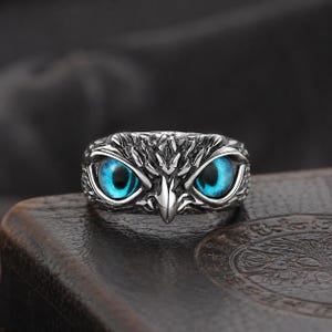 May include: A silver-toned ring featuring an owl design. The ring showcases the owl's face with bright blue eyes. The ring has a detailed, textured surface, and the owl's eyes are the focal point of the design.