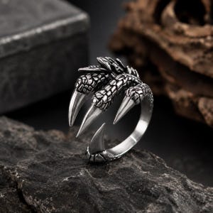 Op de afbeelding: Een zilverkleurige metalen ring met een gedetailleerd ontwerp van drie scherpe, getextureerde drakenklauwen die uit de ringband steken. Het oppervlak van de klauwen vertoont een schubpatroon, terwijl de punten glad en gepolijst zijn.