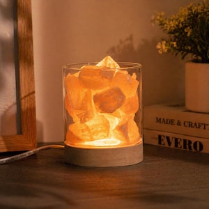 Może przedstawiać: Lampa solna himalajska. Lampa ma cylindryczny szklany pojemnik wypełniony pomarańczowymi kryształami soli. Stoi na drewnianej podstawie, emitując ciepłe światło. Lampa stoi na ciemnej drewnianej powierzchni.