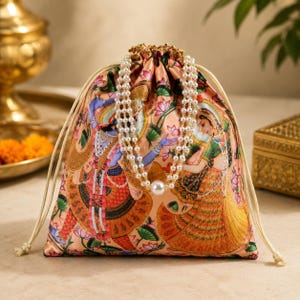 Puede incluir: Una bolsa con cordón de color melocotón con una ilustración detallada de figuras tradicionales indias. Un collar de perlas cuelga sobre la bolsa. El diseño de la bolsa incluye motivos florales y de figuras, con un cierre dorado.