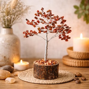 May include: A decorative tree sculpture with a silver wire trunk and branches, adorned with red-orange gemstone leaves. The tree sits in a wooden pot filled with small stones. The sculpture is on a woven mat, with candles and decorative stones nearby.