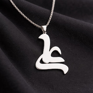 May include: A silver-colored pendant necklace featuring a stylized Arabic script design. The pendant hangs from a delicate chain, set against a dark, textured background. The necklace is a piece of jewelry.