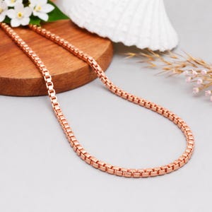 May include: A rose gold box chain necklace. The necklace is displayed on a wooden surface with white flowers and a seashell in the background. The chain is made of small, square links.