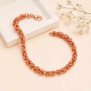 May include: A rose gold-toned chain bracelet featuring a complex Byzantine link pattern. The bracelet is constructed from numerous interlocking circular metal rings and includes a secure lobster claw clasp mechanism at one end of the chain.