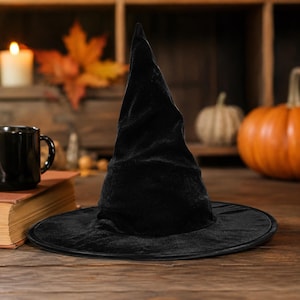 May include: A black velvet witch hat featuring a tall, pointed crown and a wide, circular brim. The fabric has a soft, textured appearance with subtle folds and creases along the conical structure, creating a dark, solid silhouette.