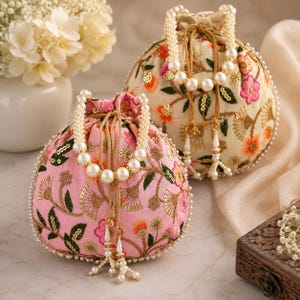 May include: Two round drawstring pouches, one in light pink and one in cream, featuring colourful floral embroidery, gold sequin accents, and white pearl bead trim. Each bag includes a pearl-beaded handle and dangling pearl-tipped tassels.