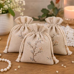 May include: Three small, beige linen drawstring pouches featuring delicate, hand-embroidered floral branch designs in soft blue, pink, and brown thread. Each pouch has a gathered top secured with matching beige cotton cord ties with tassels.
