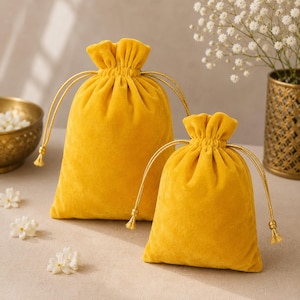 Puede incluir: Dos bolsas amarillas de terciopelo con cordón de diferentes tamaños. Las bolsas tienen cordones y borlas doradas. El fondo incluye un cuenco dorado, flores blancas y un jarrón decorativo.