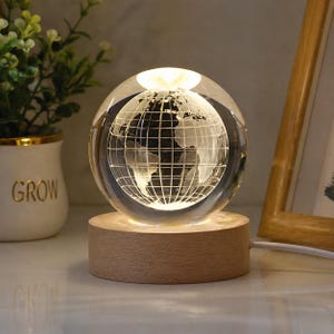 Peut inclure: Un globe en verre transparent avec une carte du monde gravée, éclairé de l'intérieur. Le globe repose sur une base en bois clair et circulaire. Une petite plante dans un pot blanc avec le mot "GROW" est visible en arrière-plan.