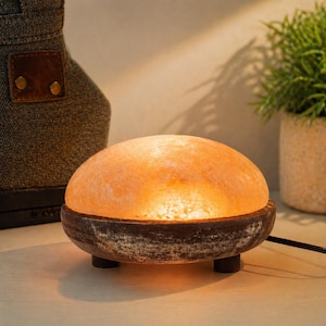 Può includere: Una lampada di sale dell'Himalaya con una calda luce arancione. La lampada è posizionata su una base marrone scuro, dall'aspetto invecchiato, con tre piccole gambe. Una piccola pianta verde in un vaso beige è sullo sfondo.