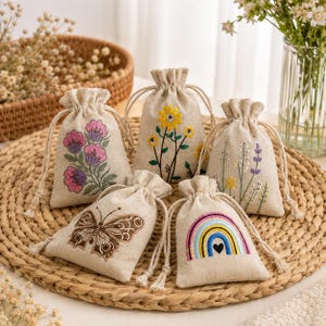 May include: Five small beige drawstring pouches made of fabric, each featuring unique embroidery. Designs include pink flowers, yellow sunflowers, lavender sprigs, a brown butterfly, and a colorful rainbow with a small black heart.
