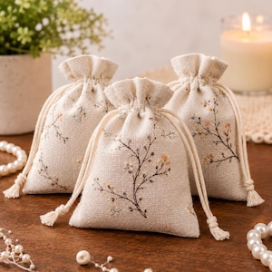 Puede incluir: Tres pequeñas bolsas con cordón de color beige hechas de tela texturizada, cada una con un delicado patrón bordado de ramas finas de color marrón con pequeños detalles florales en tonos suaves de amarillo, azul y blanco.
