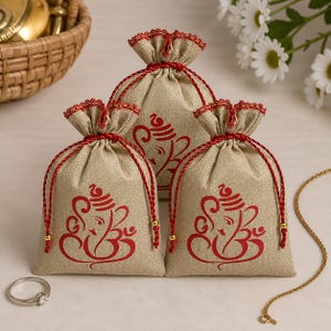 Op de afbeelding: Drie beige trekkoordzakken met een rood Ganesha-ontwerp. Elke tas heeft een rode trekkoordsluiting en afwerking. Een zilveren ring en gouden ketting staan op de voorgrond, met een rieten mand en witte bloemen op de achtergrond.