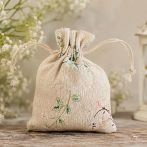 May include: A small, beige, woven fabric drawstring pouch featuring a delicate botanical print of green leaves and small pink flowers. The bag is cinched at the top with two matching beige cords, creating a gathered ruffle effect at the opening.