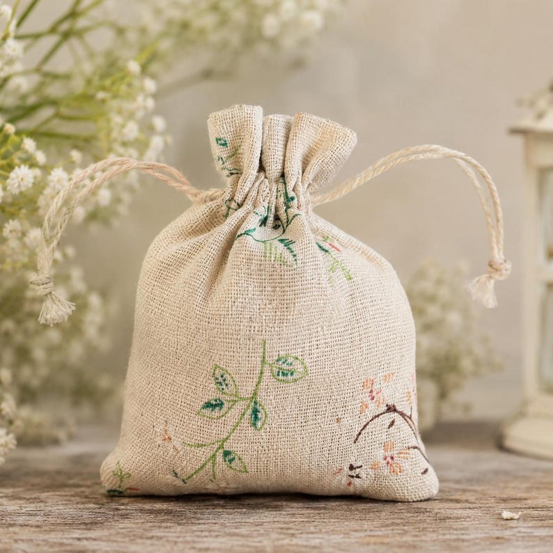 May include: A small, beige, woven fabric drawstring pouch featuring a delicate botanical print of green leaves and small pink flowers. The bag is cinched at the top with two matching beige cords, creating a gathered ruffle effect at the opening.