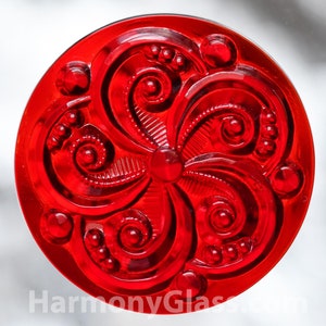 May include: A red glass button with a raised, intricate floral design. The button has a shiny, glossy finish.
