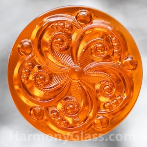 May include: Orange glass button with a swirling, floral design. The button has a raised, textured surface and a glossy finish.