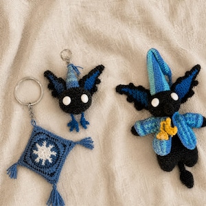 Puede incluir: Tres artículos hechos a mano con ganchillo: dos figuras de pájaros negros y azules y un cuadrado azul con un diseño de estrella blanca. Las figuras de pájaros tienen ojos blancos y alas azules. Una figura tiene un llavero adjunto.