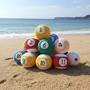 May include: A pile of crocheted snooker balls, each with a different number, on a sandy beach. The balls are in various colours, including yellow, red, green, pink, and blue. The sea and sky are visible in the background.