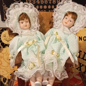 May include: Two porcelain dolls with auburn hair, wearing matching light green polka-dot dresses with white lace bonnets and collars, sitting together in a small wooden rocking chair. The dolls feature painted facial details and white shoes.