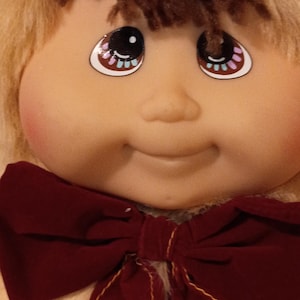 May include: A doll with a tan vinyl face, rosy cheeks, and brown yarn hair partially covered by a tan faux fur hood. The doll features large brown eyes with white highlights and wears a dark maroon velvet bow tie with yellow stitching.