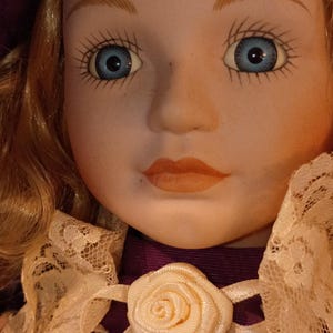 Könnte beinhalten: Eine Porzellanpuppe mit blondem Haar, blauen Augen und orangefarbenen Lippen, die einen violetten Hut und ein violettes Kleid mit einem cremefarbenen Spitzenkragen trägt. Das Kleid ist mit zwei cremefarbenen Stoffrosen verziert.