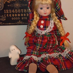 May include: A vintage doll with blonde plaits, dressed in a red plaid dress and bonnet. A small white ghost figurine is next to the doll. A Ouija board is in the background.