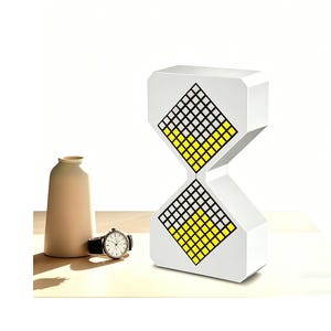 May include: A white hourglass-shaped object featuring two diamond-shaped displays with a grid of yellow and grey squares. Beside it sits a beige ceramic vase and a wristwatch with a black leather strap and a white face with black hands.