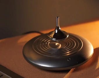 May include: A levitating top with a polished chrome finish, hovering above a circular base with concentric ring patterns. The base is a dark metallic color, and the top appears to be spinning. The background is a warm, neutral tone.