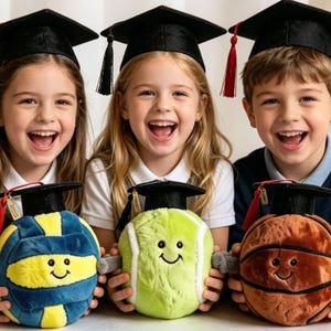 May include: Three children wearing black graduation caps with tassels, each holding a plush sports ball toy. The toys include a blue and yellow volleyball, a lime green tennis ball, and an orange basketball, all featuring smiling faces.