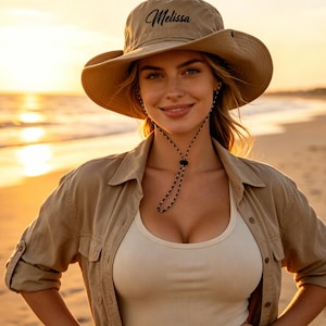 Puede incluir: Un sombrero de sol color canela de ala ancha con el nombre Melissa bordado en letra cursiva negra en la parte delantera. El sombrero incluye una correa ajustable para la barbilla con un patrón en blanco y negro y un cierre.